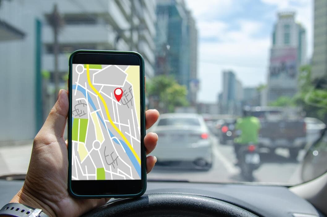 A phone showing a navigation map with a location pin, held up in front of a car's steering wheel on a city street
